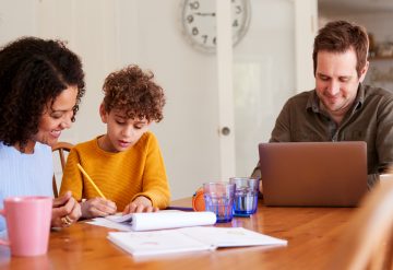 Father Works On Laptop As Mother Helps Son With Homework On Kitchen Table